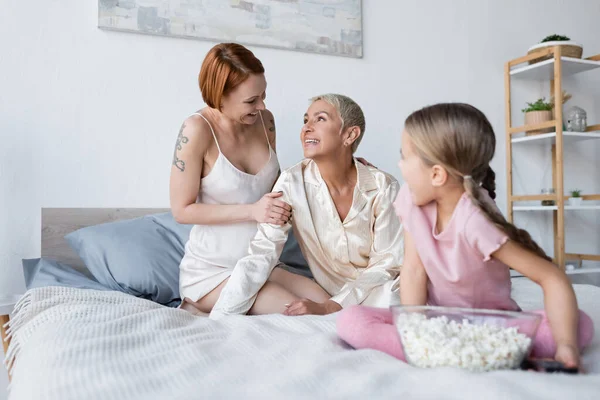 Smiling woman touching girlfriend near blurred kid with popcorn on bed — Stock Photo