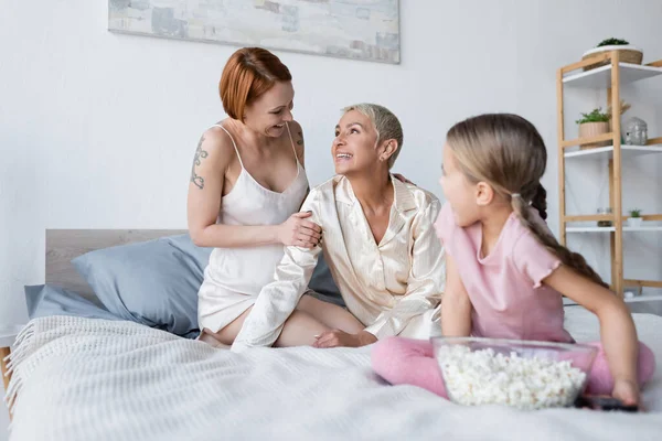 Mujer sonriente tocando novia cerca de niño borroso con palomitas de maíz en la cama - foto de stock