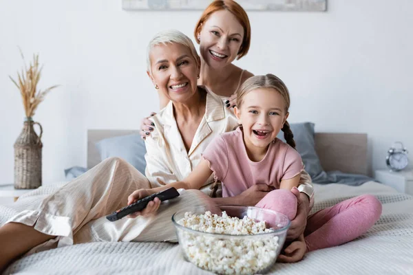 Ragazza positiva tenendo telecomando vicino popcorn e madri sul letto — Foto stock