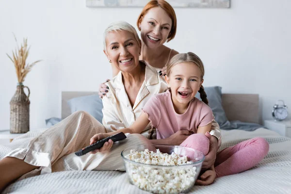 Menina positiva segurando controlador remoto perto de pipocas e mães na cama — Fotografia de Stock