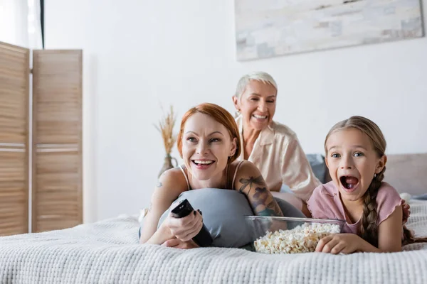 Aufgeregtes Kind schaut Film in der Nähe von Popcorn und Müttern im Bett — Stockfoto