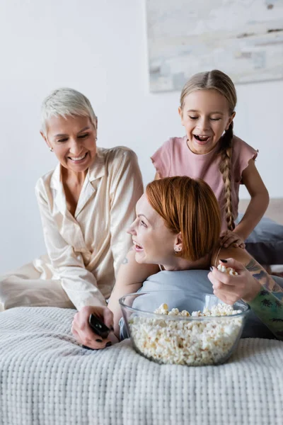 Sorridente donna lesbica che tiene popcorn e telecomando vicino a bambino e fidanzata sul letto — Foto stock