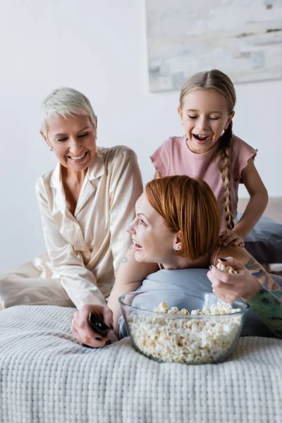 Sonriente lesbiana sosteniendo palomitas de maíz y mando a distancia cerca de niño y novia en la cama - foto de stock