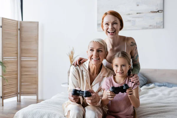 KYIV, UKRAINE - DECEMBER 8, 2021: Positive lesbian woman hugging girlfriend and daughter with joysticks on bed at home — Stock Photo