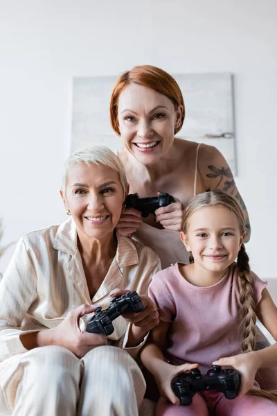KYIV, UCRAINA - 8 DICEMBRE 2021: Felice coppia lesbica e figlia adottiva che tiene i joystick e guarda la fotocamera a casa — Foto stock