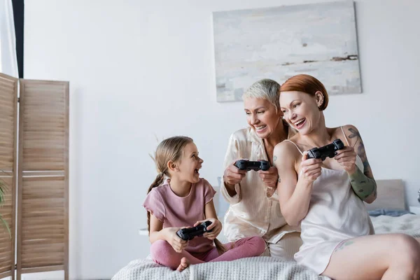 KYIV, UCRAINA - 8 DICEMBRE 2021: Coppia lesbica felice e figlia adottiva che gioca ai videogiochi in camera da letto — Foto stock