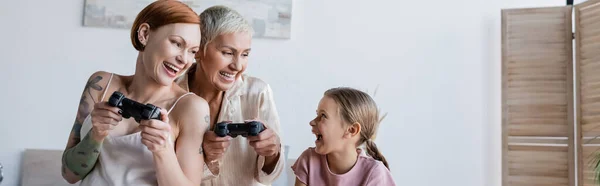 KYIV, UCRANIA - 8 DE DICIEMBRE DE 2021: Niño positivo mirando a las madres con joysticks en casa, pancarta - foto de stock