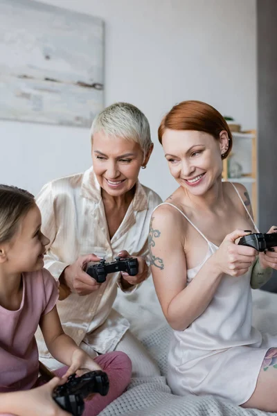 KYIV, UKRAINE - DECEMBER 8, 2021: Smiling lesbian couple holding joysticks near adopted daughter on bed — Stock Photo
