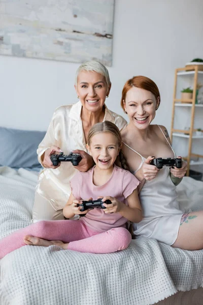 KYIV, UKRAINE - DECEMBER 8, 2021: Smiling lesbian couple and kid holding joysticks on bed — Stock Photo