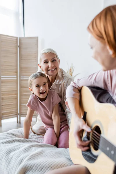 Lesbische Frau und Kind schauen verschwommene Freundin an, die im Schlafzimmer Akustikgitarre spielt — Stockfoto