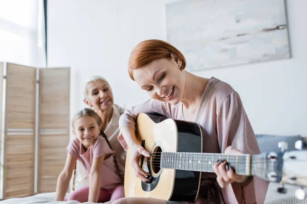 Femme lesbienne positive jouant de la guitare acoustique près de petite amie floue et enfant à la maison — Photo de stock