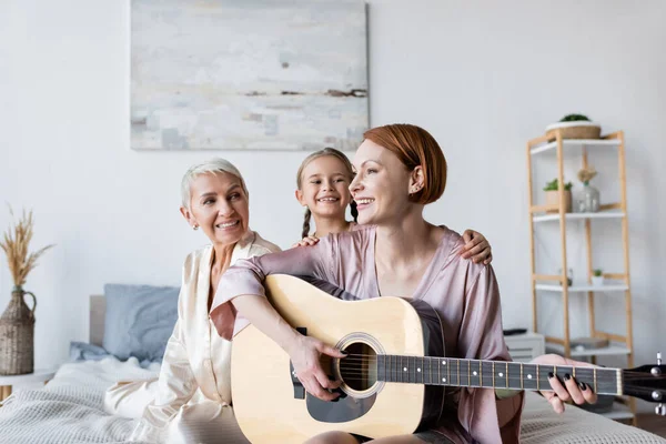 Mujer sonriente y niño mirando a su novia tocando la guitarra acústica en la cama - foto de stock