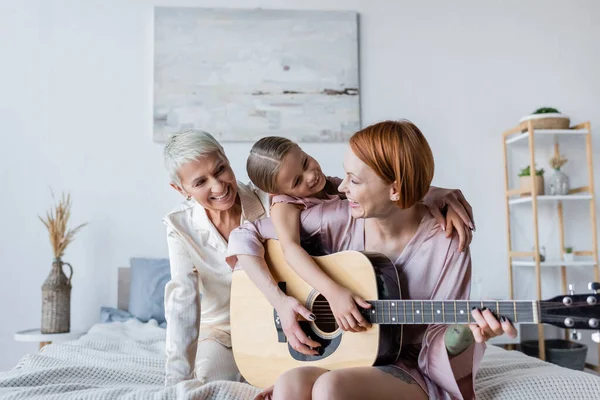 Positive woman playing acoustic guitar near adopted daughter and girlfriend on bed — Stock Photo