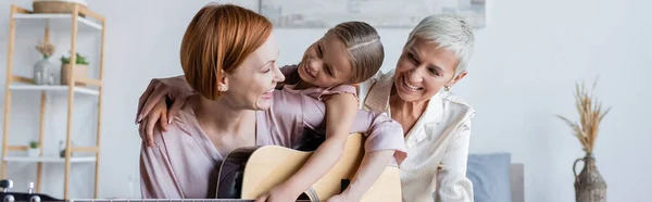 Fröhliches Kind umarmt lesbische Mutter mit Akustikgitarre im Schlafzimmer, Banner — Stockfoto