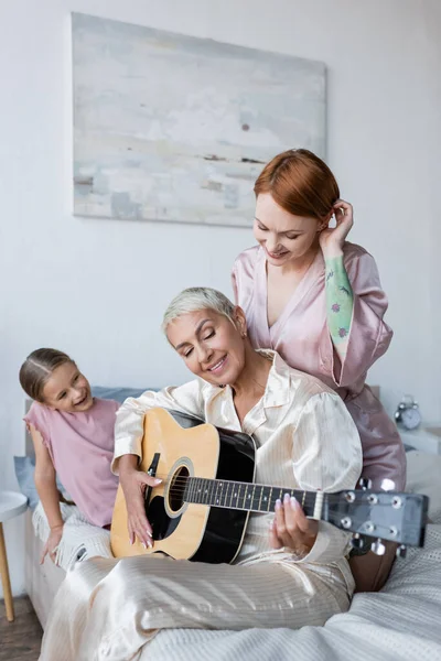 Mujer sonriente mirando a su novia con guitarra acústica cerca de su hija en la cama - foto de stock
