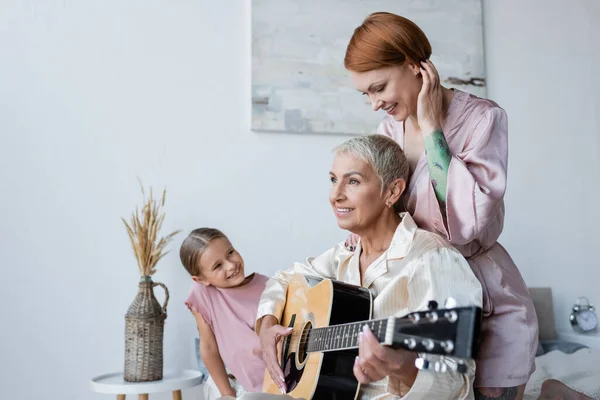 Lesbian woman playing acoustic guitar near adopted daughter and girlfriend on bed — Stock Photo