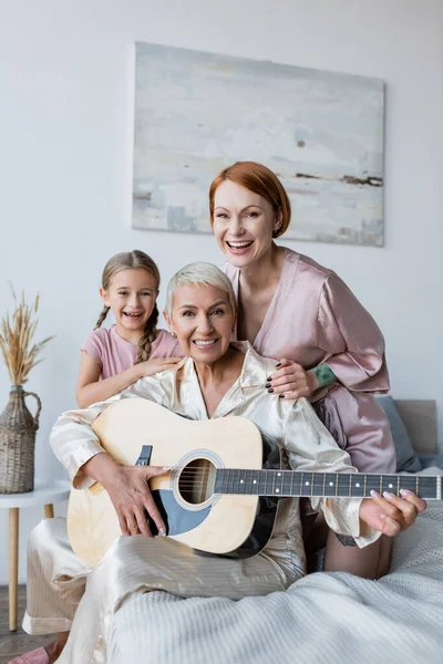 Positiva coppia lesbica con chitarra acustica e figlia guardando la fotocamera sul letto a casa — Foto stock