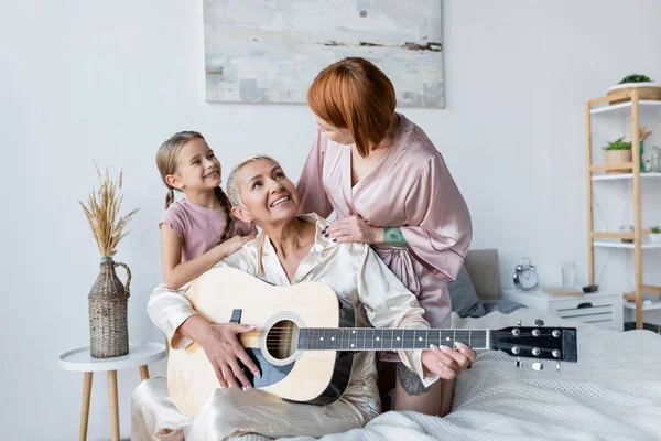 Fröhliche lesbische Frau spielt Akustikgitarre neben Kind und Freundin im Schlafzimmer — Stockfoto