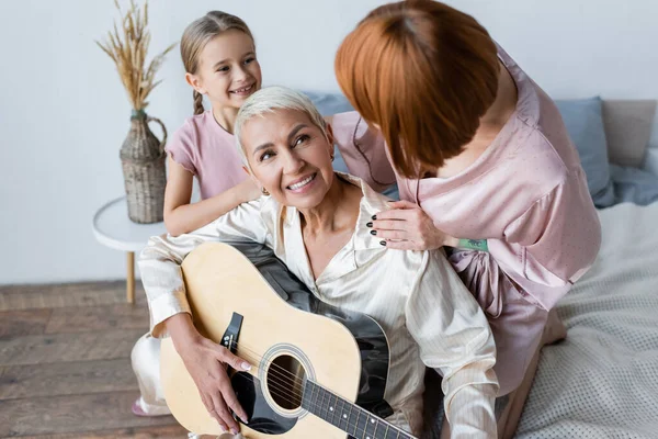 Femme souriante jouant de la guitare acoustique près de la fille adoptée et la petite amie dans la chambre — Photo de stock