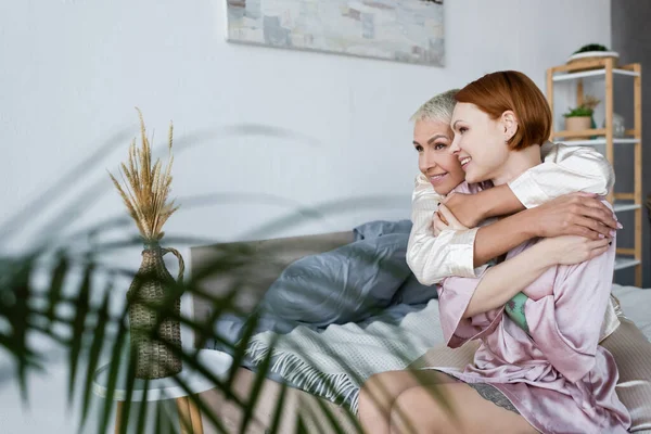 Glückliches lesbisches Paar umarmt sich auf dem Bett in der Nähe von Pflanzen zu Hause — Stockfoto