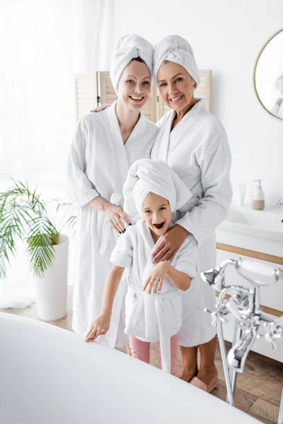 Positives lesbisches Paar in Bademänteln umarmt Adoptivtochter nahe Badewanne im Badezimmer — Stockfoto