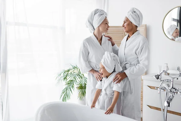 Smiling lesbian couple standing with adopted daughter in bathrobe near bathtub at home — Stock Photo