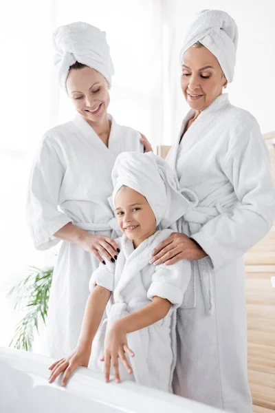 Lesbian women in bathrobes standing near daughter and bathtub at home — Stock Photo
