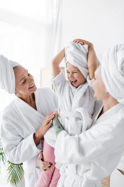 Happy lesbian women in bathrobes holding positive adopted daughter in bathroom — Stock Photo