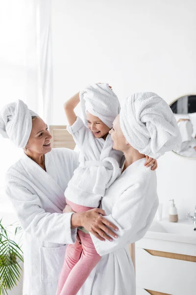 Feliz pareja de lesbianas sosteniendo a su hija en toalla y albornoz en el baño - foto de stock