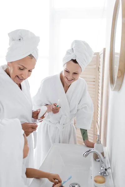 Happy lesbian couple in bathrobes holding toothpaste and toothbrushes near sink and daughter in bathroom — Stock Photo