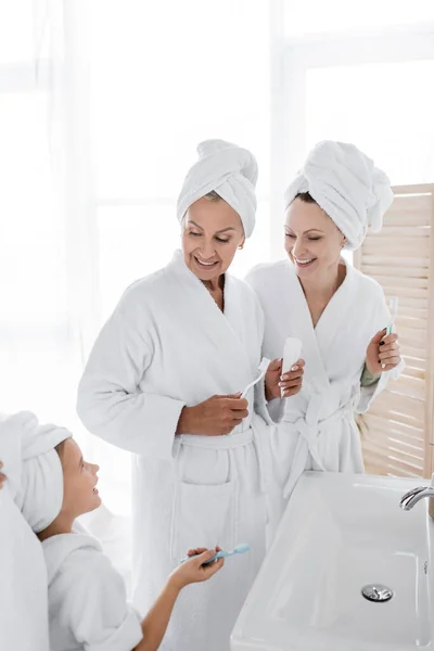 Smiling lesbian women in bathrobes holding toothbrushes near blurred adopted daughter in towel — Stock Photo