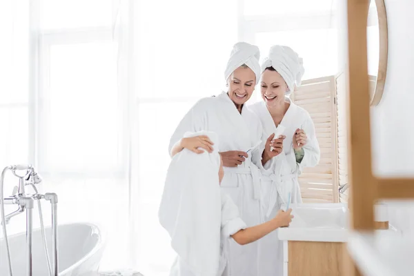 Positivos padres lesbianas en batas de baño sosteniendo cepillos de dientes cerca de hija adoptada en el baño - foto de stock