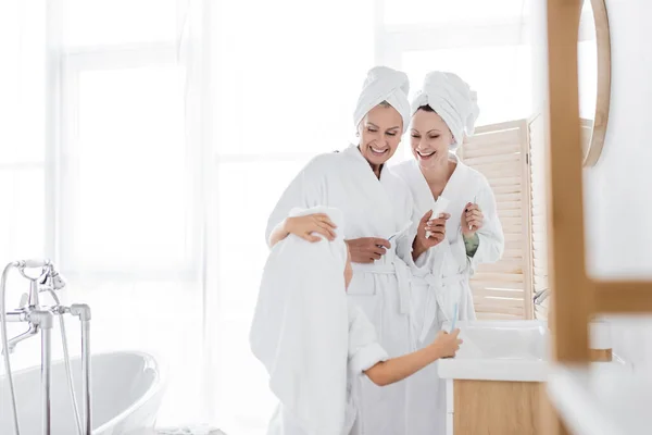 Parents lesbiennes positifs en peignoirs tenant des brosses à dents près de la fille adoptée dans la salle de bain — Photo de stock