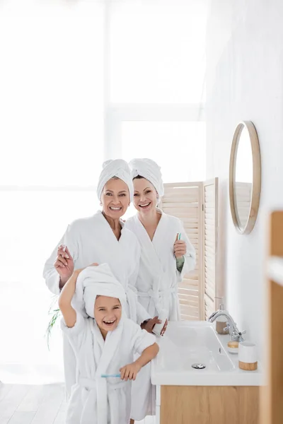 Cheerful lesbian parents in bathrobes holding toothbrushes near daughter in bathroom — Stock Photo