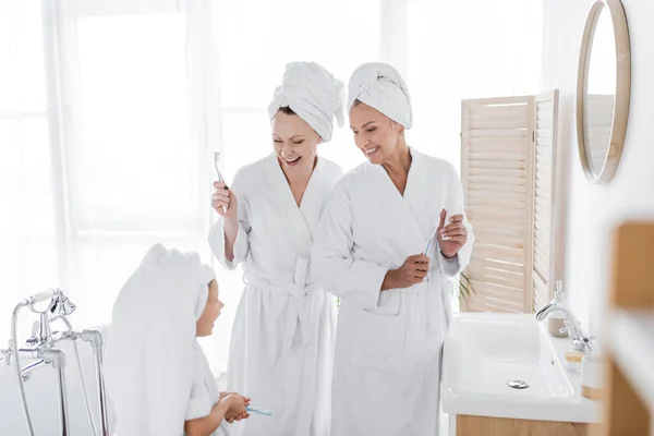 Smiling kid in towel holding toothbrush near mothers in bathroom — Stock Photo