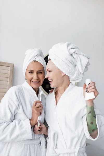 Pareja lésbica positiva en toallas y albornoces sosteniendo cepillos de dientes en el baño - foto de stock