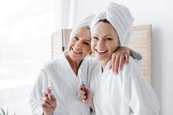Smiling lesbian woman in bathrobe hugging girlfriend with toothbrush at home — Stock Photo