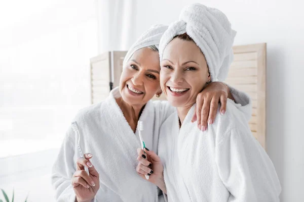 Femme lesbienne souriante en peignoir étreignant sa petite amie avec une brosse à dents à la maison — Photo de stock