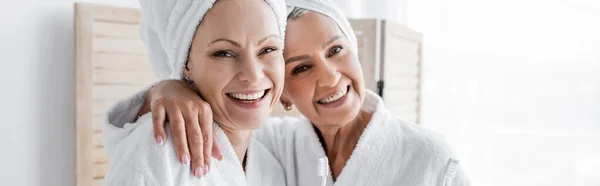 Femme souriante en peignoir et serviette étreignant petite amie avec brosse à dents à la maison, bannière — Photo de stock