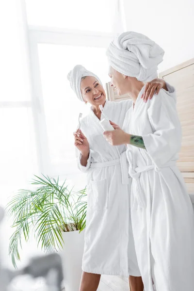 Smiling lesbian couple in bathrobes holding toothbrushes at home — Stock Photo