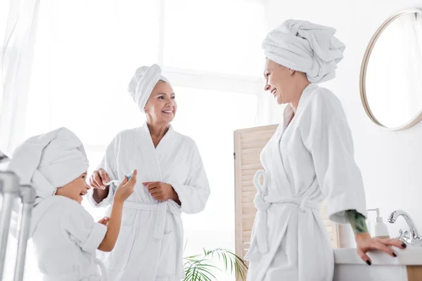 Smiling kid in bathrobe holding toothpaste near mothers in bathroom — Stock Photo