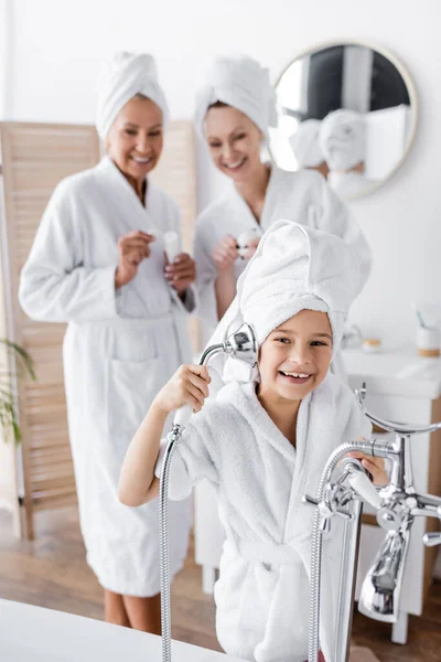 Cheerful kid in bathrobe holding shower head near blurred moms at home — Stock Photo