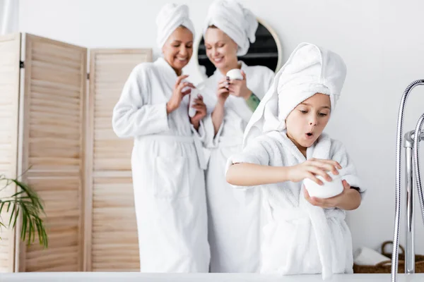 Erstauntes Mädchen im Bademantel hält Kosmetikcreme in der Nähe verschwommener Mütter im Badezimmer — Stockfoto