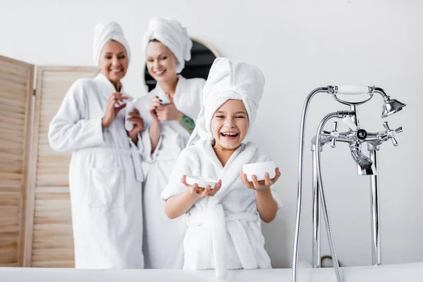 Щаслива дівчина в рушнику і халаті тримає косметичний крем біля розмитих матерів у ванній — стокове фото