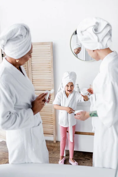 Весела дитина в халаті тримає косметичний крем біля розмитих матерів у ванній — стокове фото