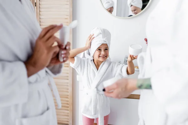 Criança positiva em toalha e roupão segurando creme cosmético perto das mães no banheiro — Fotografia de Stock