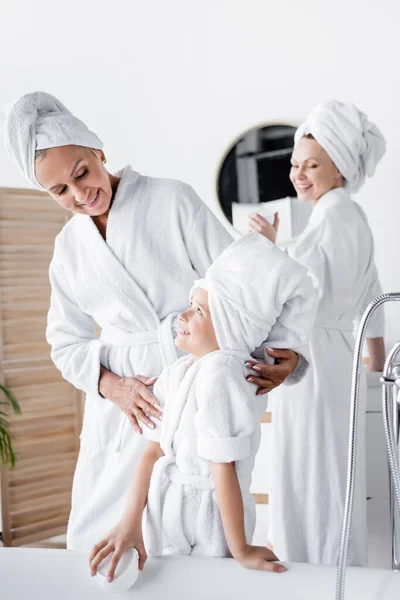 Mujer sonriente en albornoz abrazando a la hija cerca de novia borrosa en el baño - foto de stock