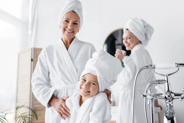 Sonriente niño en toalla y albornoz mirando a la cámara cerca de mamá en casa - foto de stock