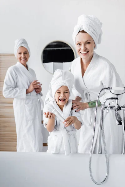 Позитивна жінка в халаті обіймає дочку косметичним кремом у ванній — стокове фото