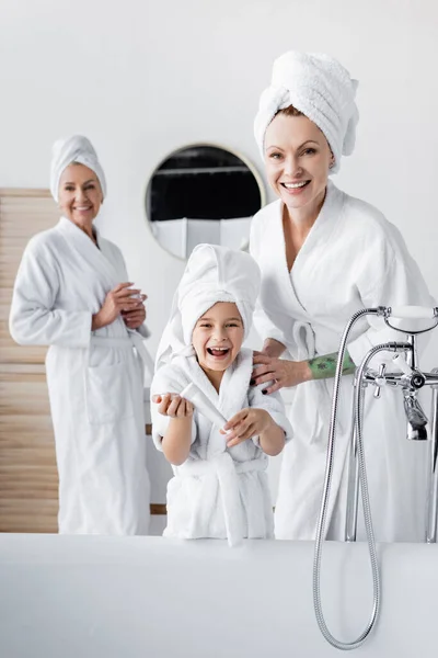 Mujer positiva en albornoz abrazando hija con crema cosmética en el baño - foto de stock